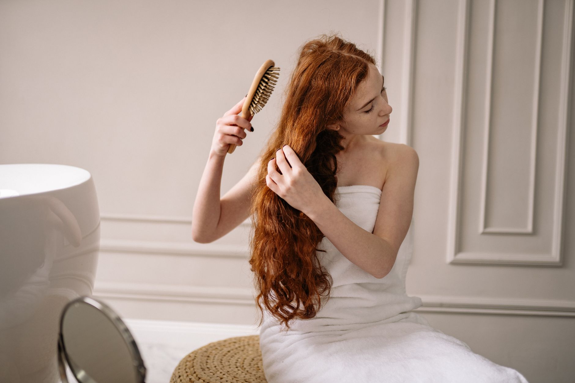 A small ceramic bowl of melted coconut oil next to a wide-tooth comb, a folded towel, and a hair clip arranged on a bathroom shelf.