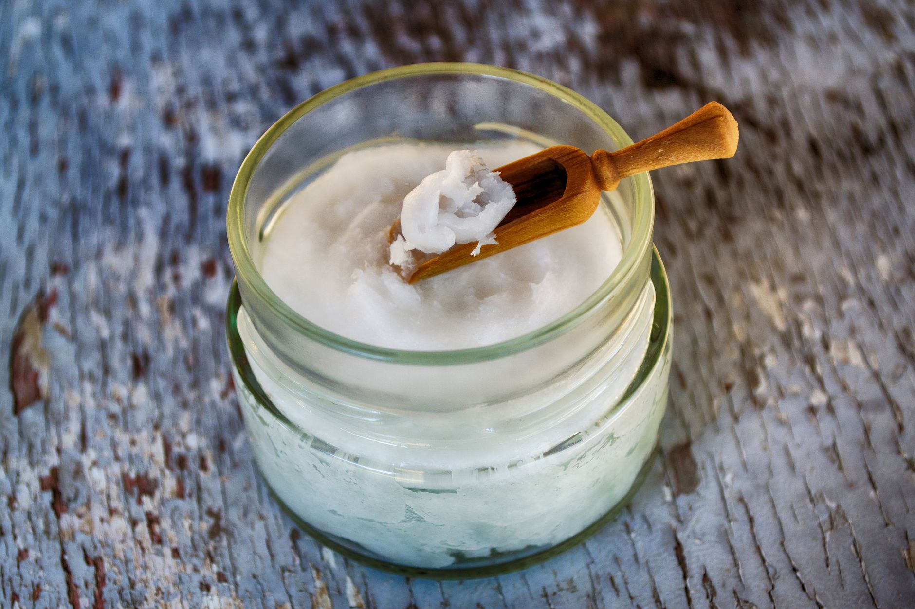 An open glass jar of solid white coconut oil beside a small bottle of clear liquid oil and a wooden spoon on a light countertop.